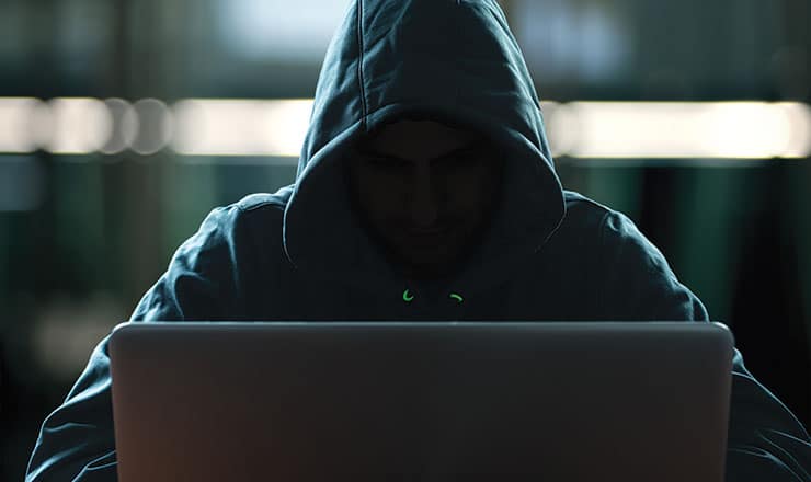 A sinister image of a hooded person sitting at a keyboard, indicating scams and fraud
