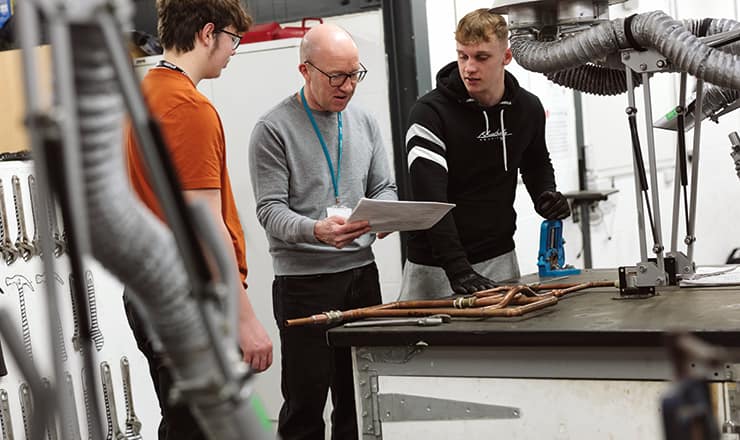 An older man standing between two younger males, with pipework on a bench in front, and holding a document, indicating teaching plumbing apprentices